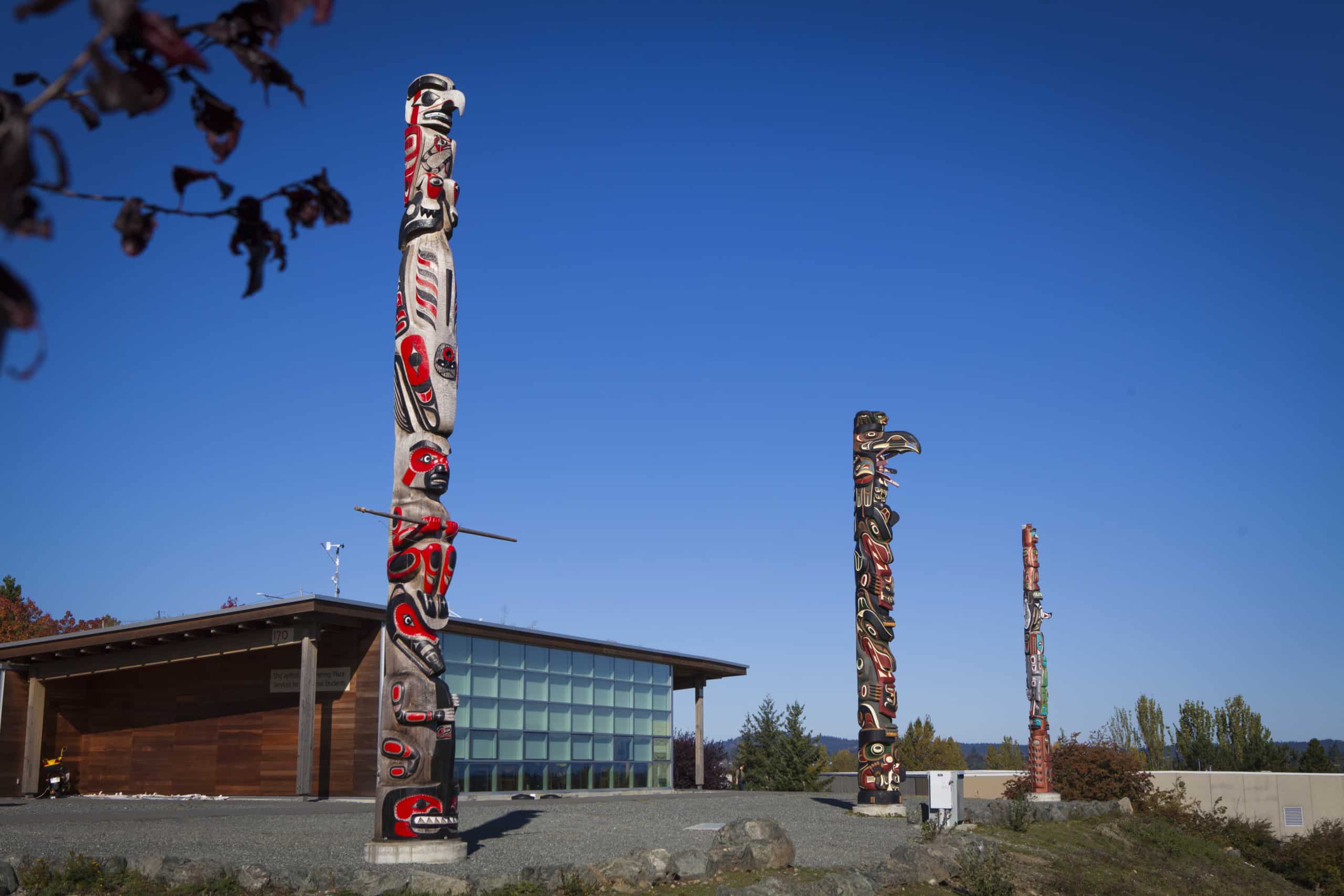 Three totem poles stand out against a very blue sky. A squat building with a flat roof and a glass wall stand behind.