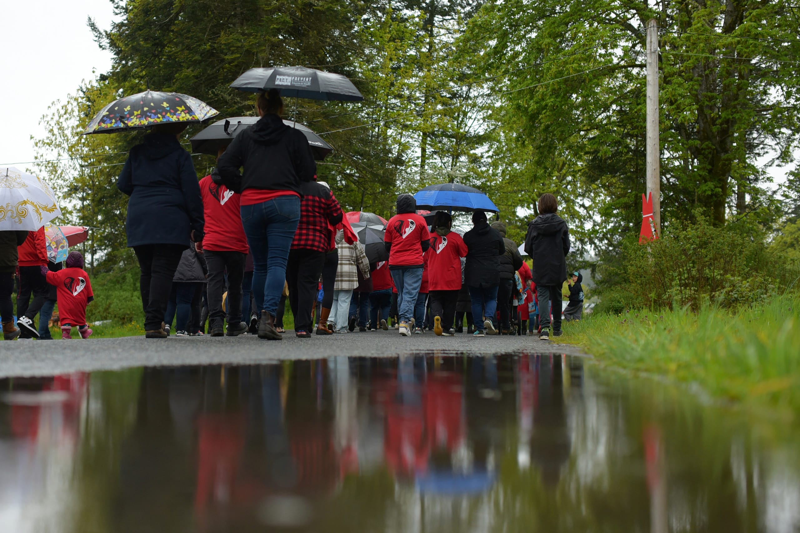 A crowd of people who are each wearing an item of red clothing in honour of MMIWG2S walk away from the camera, along a path beside a small, shallow body of water. It is raining and overcast outside, and some are carrying umbrellas.