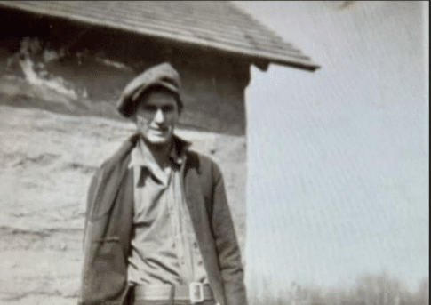 A young nehiyaw man, Alphonse Little Poplar, stands in front of a wooden building on the prairie.