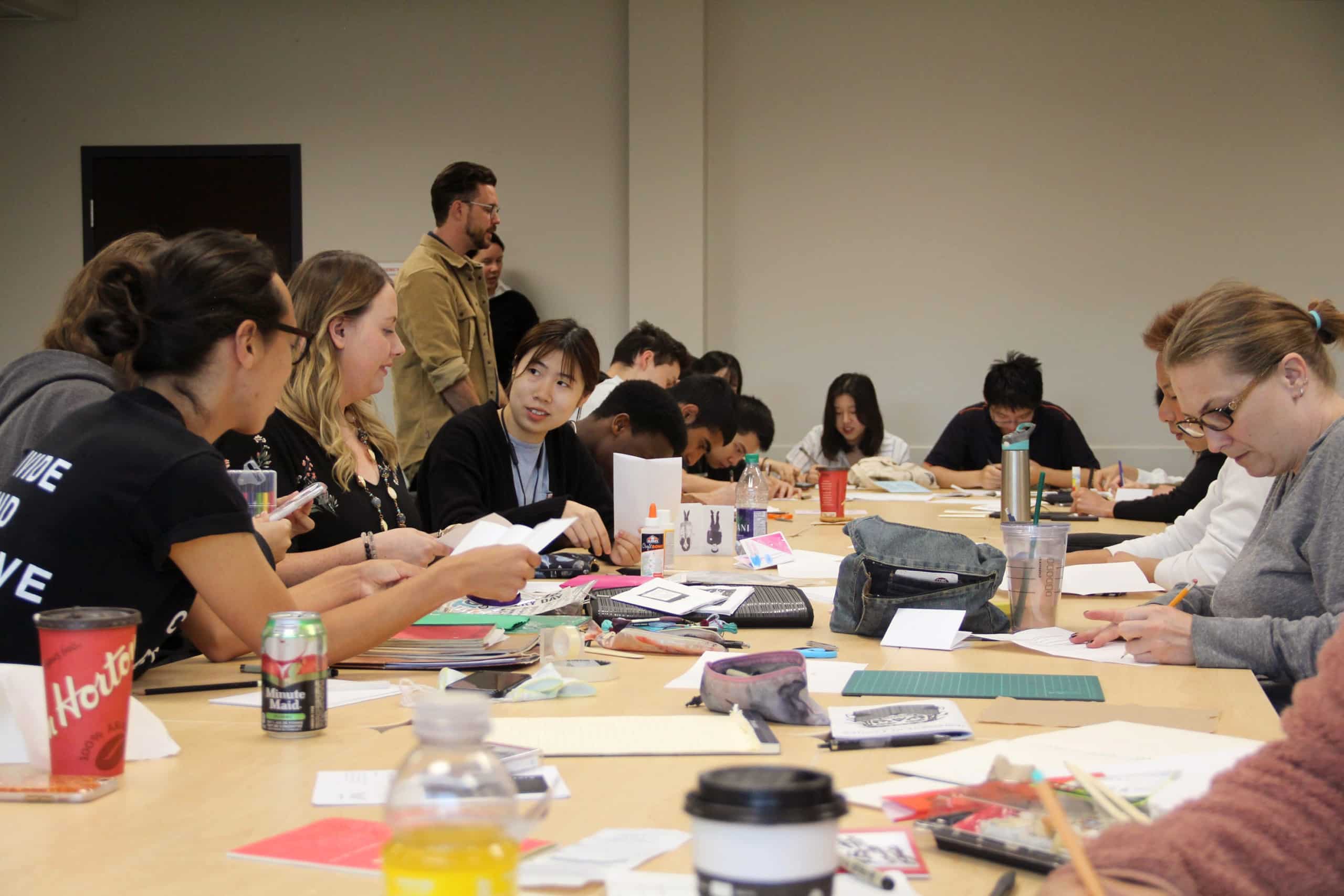 A group of students are seen around a large table,participate in a ‘zine’ making workshop, led by Whess Harman during the 2019 Indigenous Art Intensive.