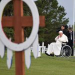 A wooden grave marker, resembling a cross encircled with a Medicine Wheel, is visible in the foreground. Pope Francis sits in a wheelchair on a green field overlooking the graveyard.