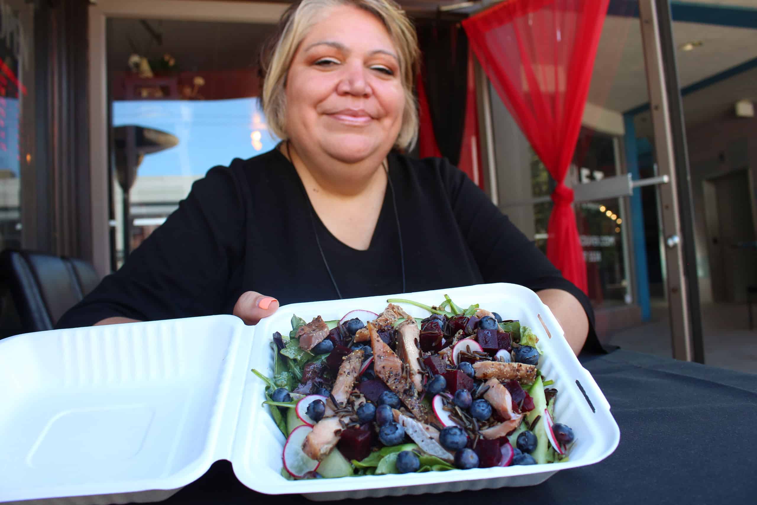 A woman holds a takeout box containing a salad with blueberries and salmon
