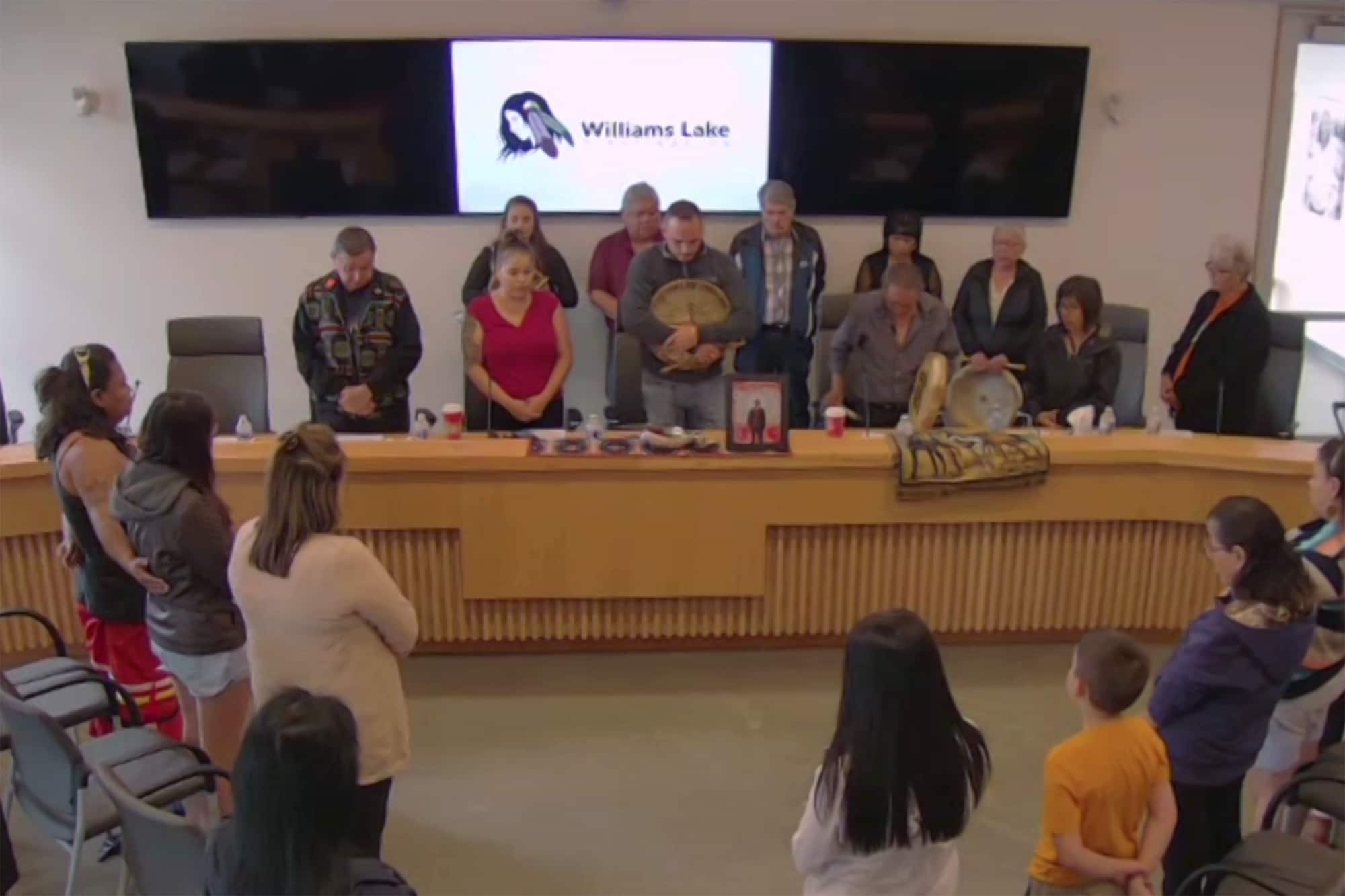 Ten people are seen standing behind a desk in the Williams Lake First Nation's office. More people are standing up from their seats to face the leaders at the front of the room.