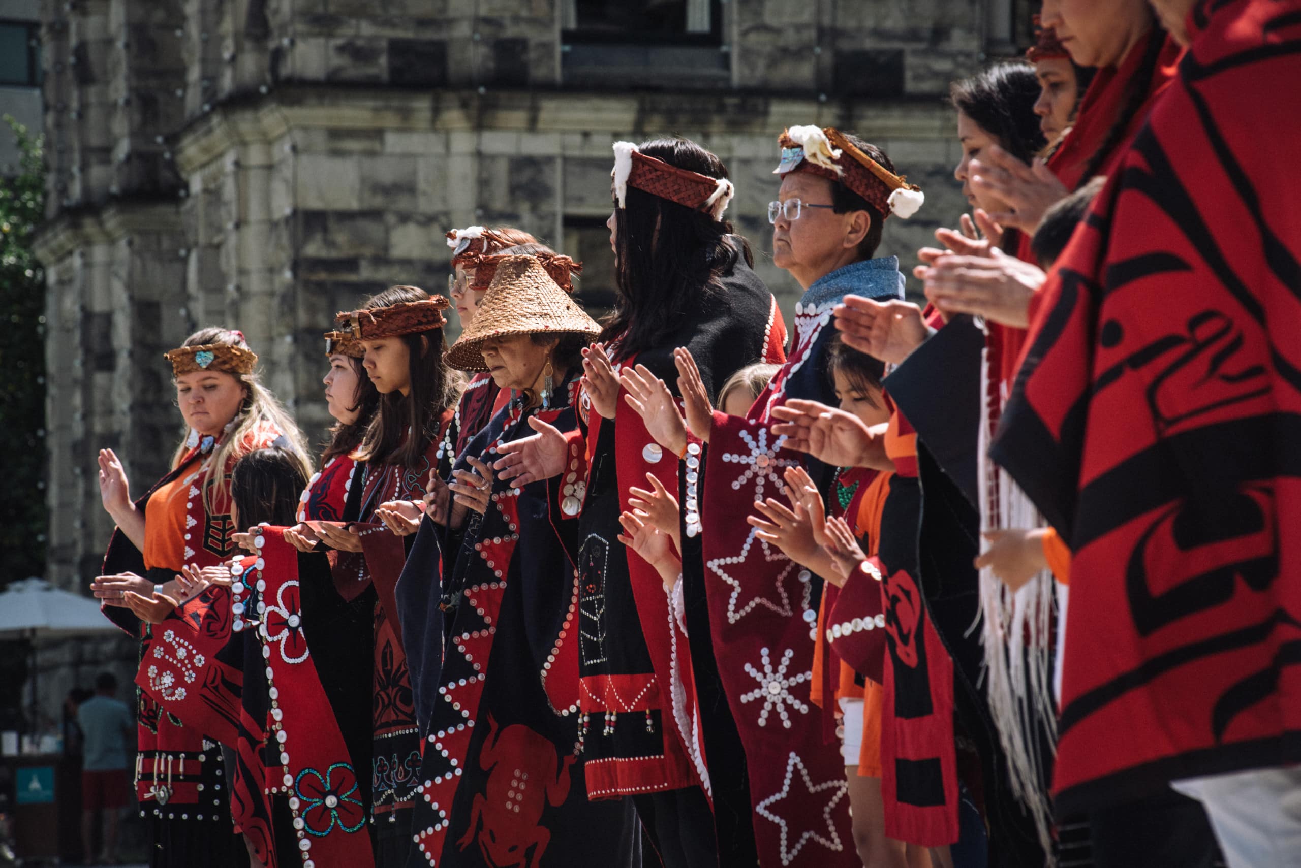 A group of Indigenous people clad in black and red button blankets and traditional cedar hats stand together in ceremony.