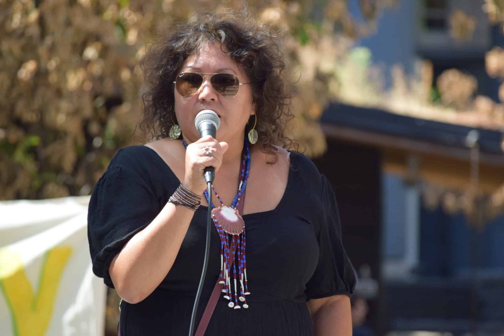 Charlene Aleck of Tsleil-Waututh Nation stands speaking into a microphone, adorned in abalone shell earrings and a beaded an shell necklace.