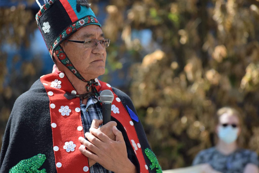 Wet’suwet’en Hereditary Chief Namoks stands with a microphone held to his chest. He is wearing a red and black button blanket and a matching felt hat.