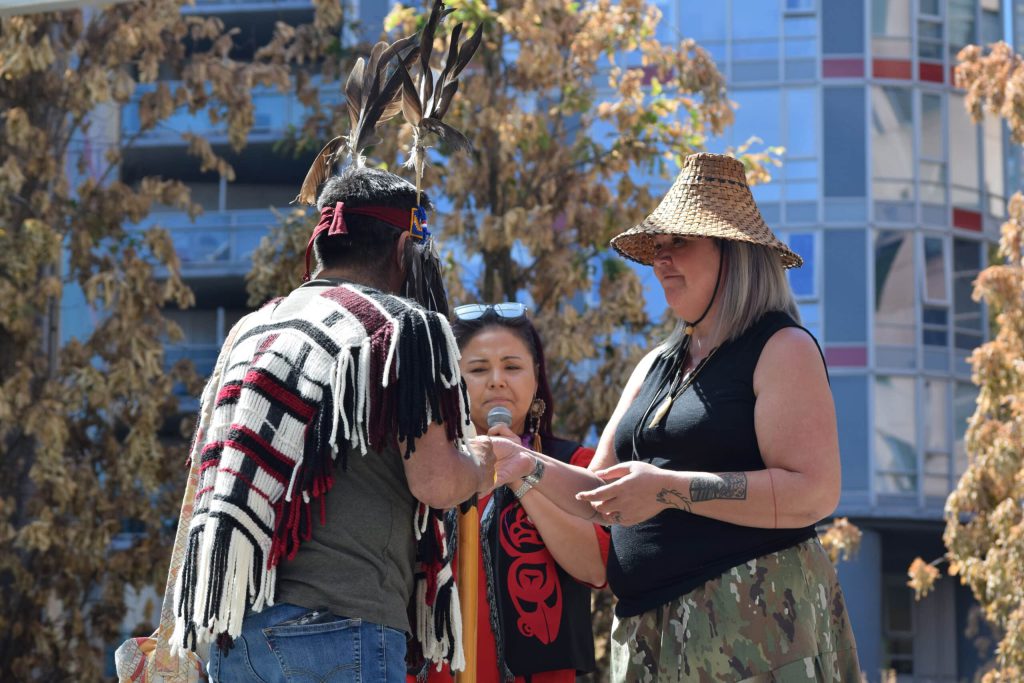 Robert Nahanee of Squamish Nation is wearing regalia of a woven wool poncho and headband, is gifted salmon from Wet’suwet’en homelands by Jennifer Wickham, who is wearing a woven cedar hat and black shirt.