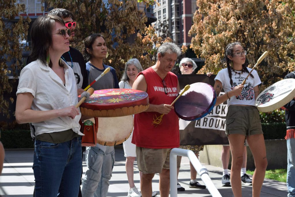 Five Indigenous people stand in a semi-circle side by side, singing and drumming together in "Vancouver".