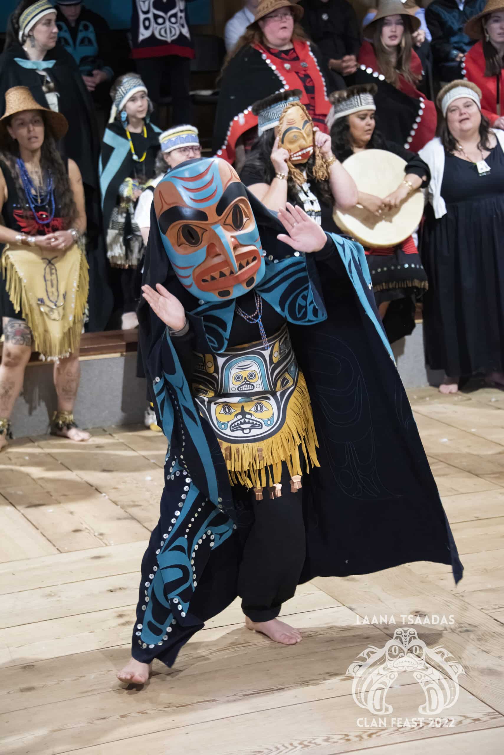 A Haida woman is wearing a carved wooden mask with a dogfish design, she is dancing on a wooden floor in the longhouse while guests watch in the background.
