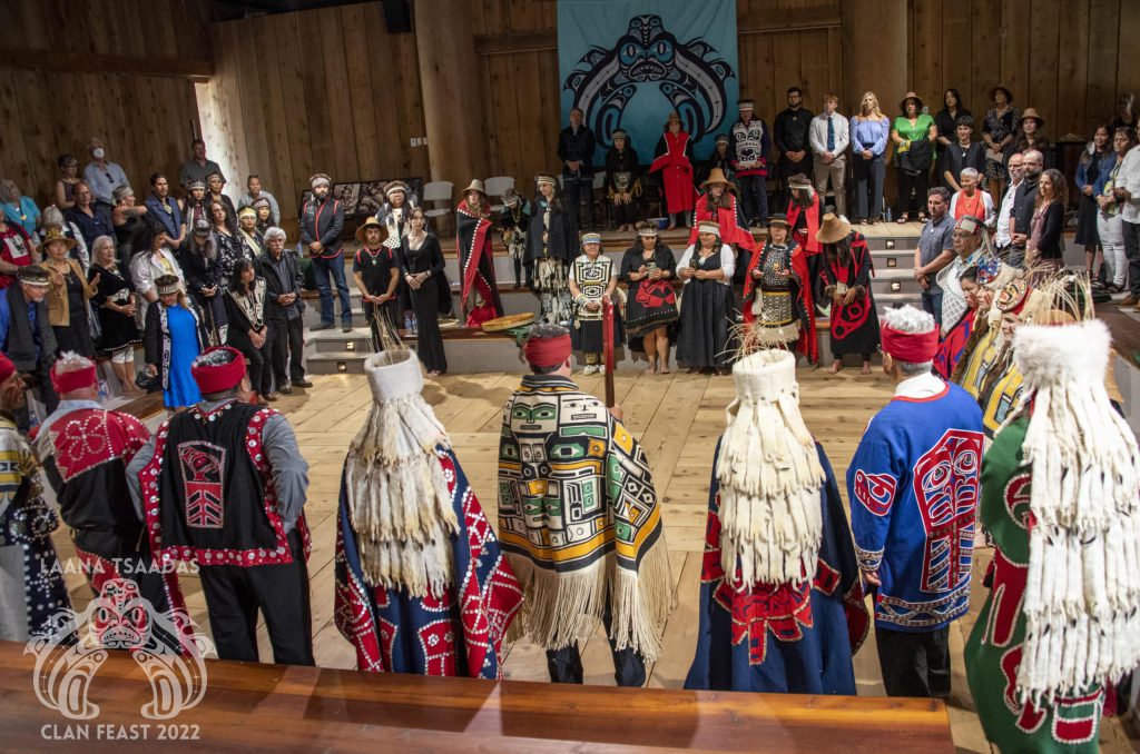 Dozens of people stand in a cedar wood longhouse along the panelled walls. The two families of the marrying couple are standing across the floor, facing each other. Most people are wearing regalia, including red and black button blankets and vests. Head pieces that are wrapped around their heads, chilkat woven blankets, furs, and shells.