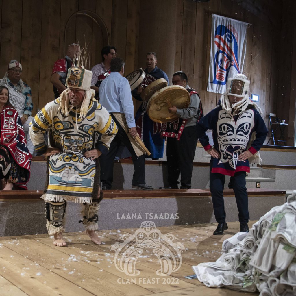 Two men dance together in their regalia and head pieces made of fur, copper, and seal whiskers. There is a group of people behind them, singing and drumming, and others watch the dance.