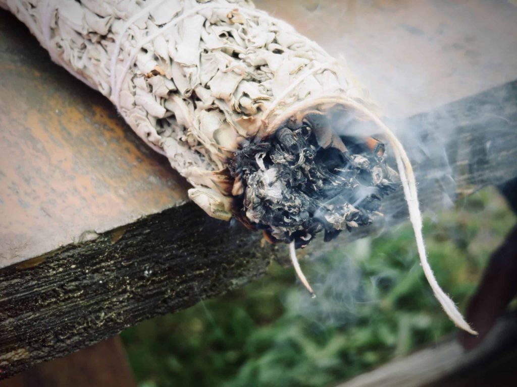 A bundle of sage is burning while placed on a table, it's smudge smoke is wafting up from it.