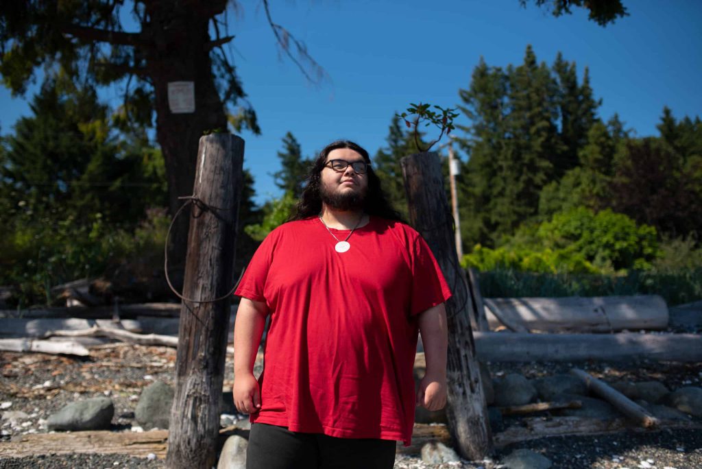 Isaiah Harris stands on the beach, against a backdrop of the hightide line marked by driftwood and stones. He is wearing a red t-shirt and black framed glasses in the sunshine.