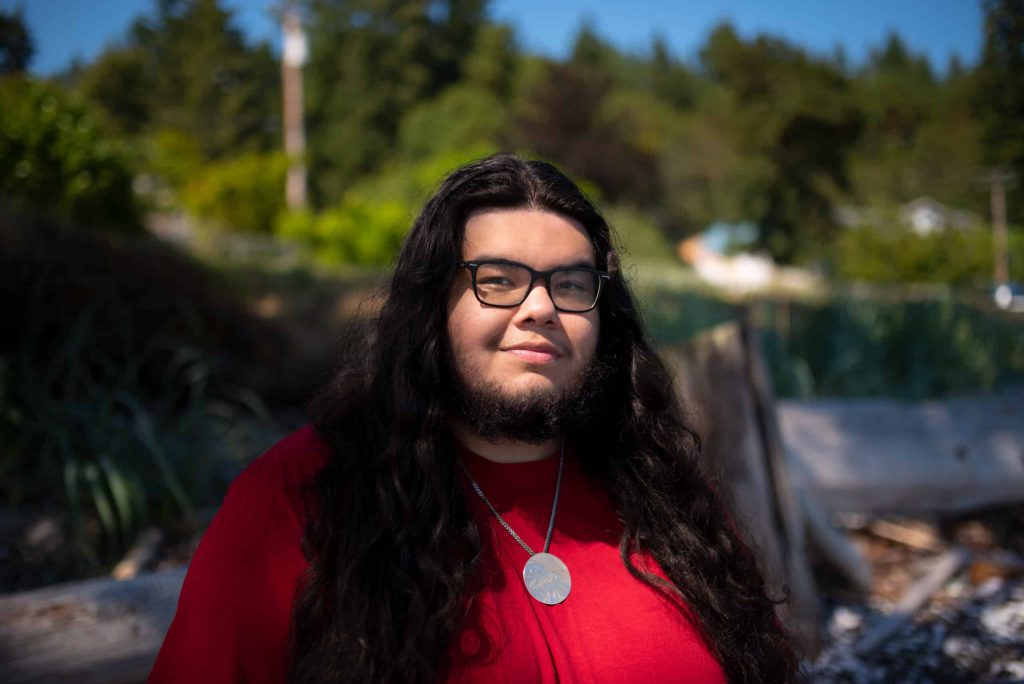Isaiah Harris is wearing a red t-shirt, with his long black hair framing his face and going past his chest. He is wearing glasses and standing on the beach with water behind him in the sunshine.