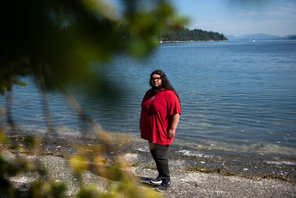 Isaiah Harris stands on the beach in a bright red t-shirt and black jeans, facing away from the ocean behind him. 