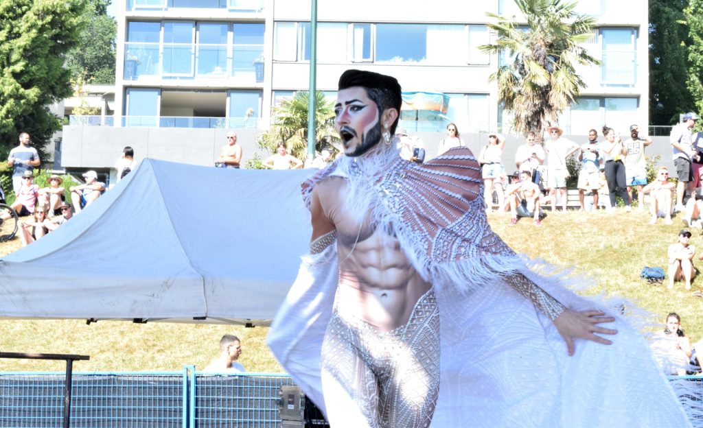A person with short hair and a beard performs on stage wearing a white cape