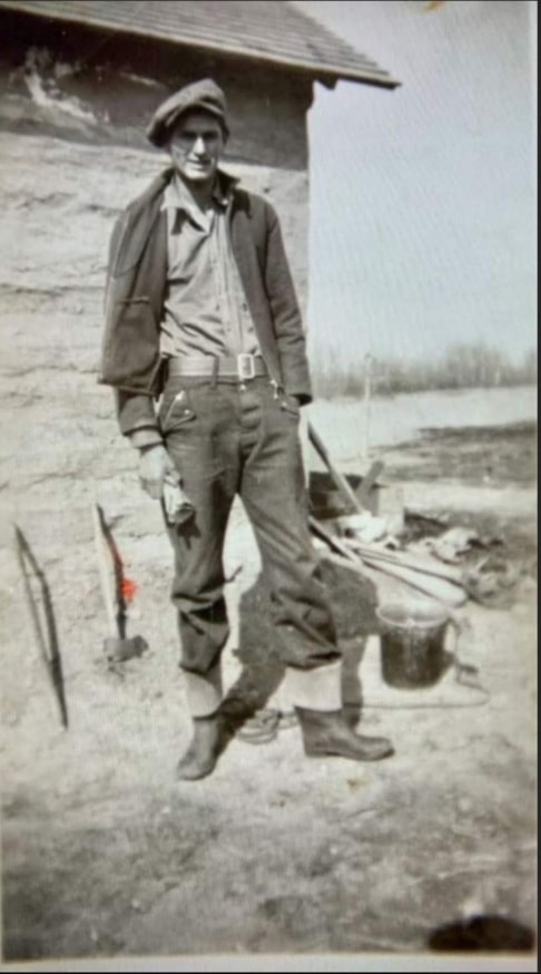 A young nehiyaw man stands in front of a wooden building on the prairie.