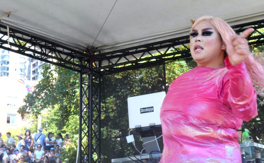 A person with long pink hair and a shiny tight pink dress with long sleeves dances on stage