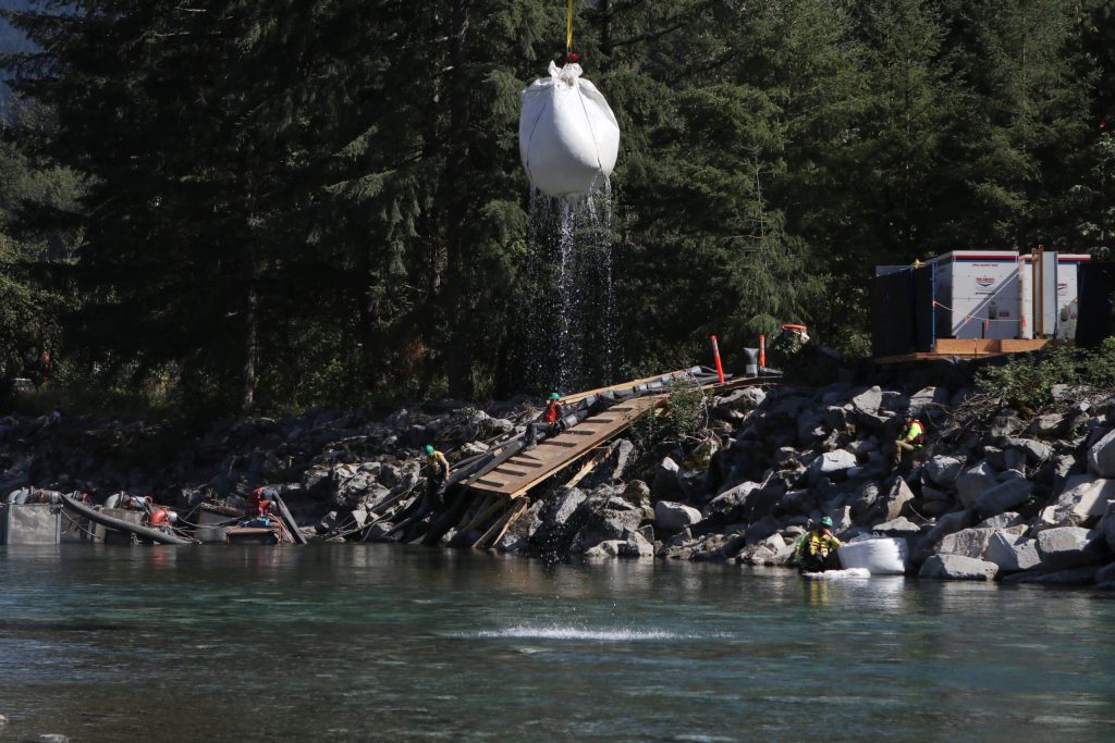 A huge white sandbag is seen airlifted out of the river with a crane with water streaing down from it and workers watching from the riverbank.