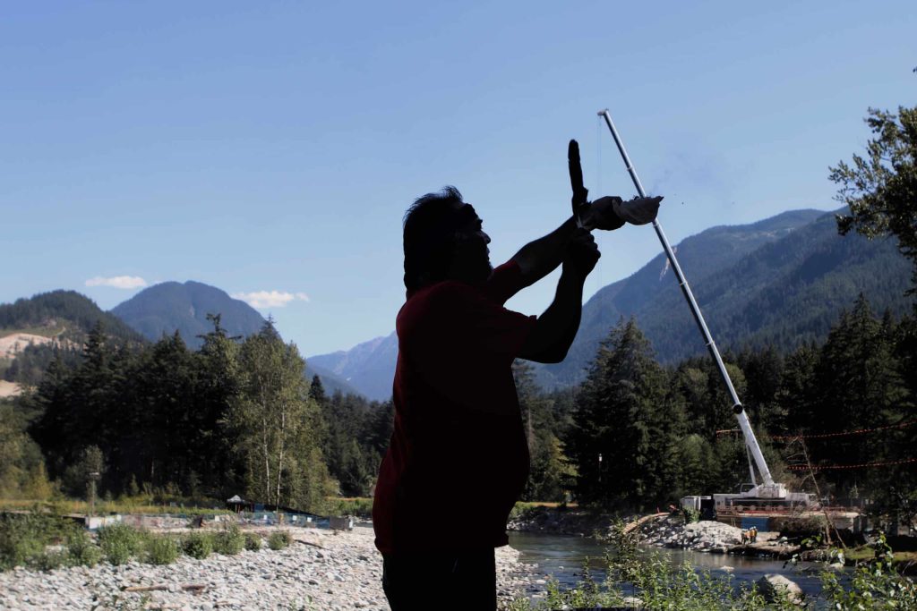 Tim Henry, a Vuntut Gwitchin and Tlingit man smudges the Kw'ikw'iya:la River within S'olh Temexw during a ceremony, while workers with the Trans Mountain Expansion (TMX) project complete the finishing touches of an installation of a pipeline under the waterway.