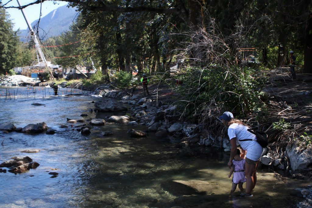 Patricia Dion, a Cree resident of “Hope, B.C.,” watches with the child of a family friend as workers with the Trans Mountain Expansion (TMX) project remove equipment from the Kw'ikw'iya:la River within S'olh Temexw. Sandbags and water pumps were used to create a dam during the trenched installation of a pipeline under the waterway.