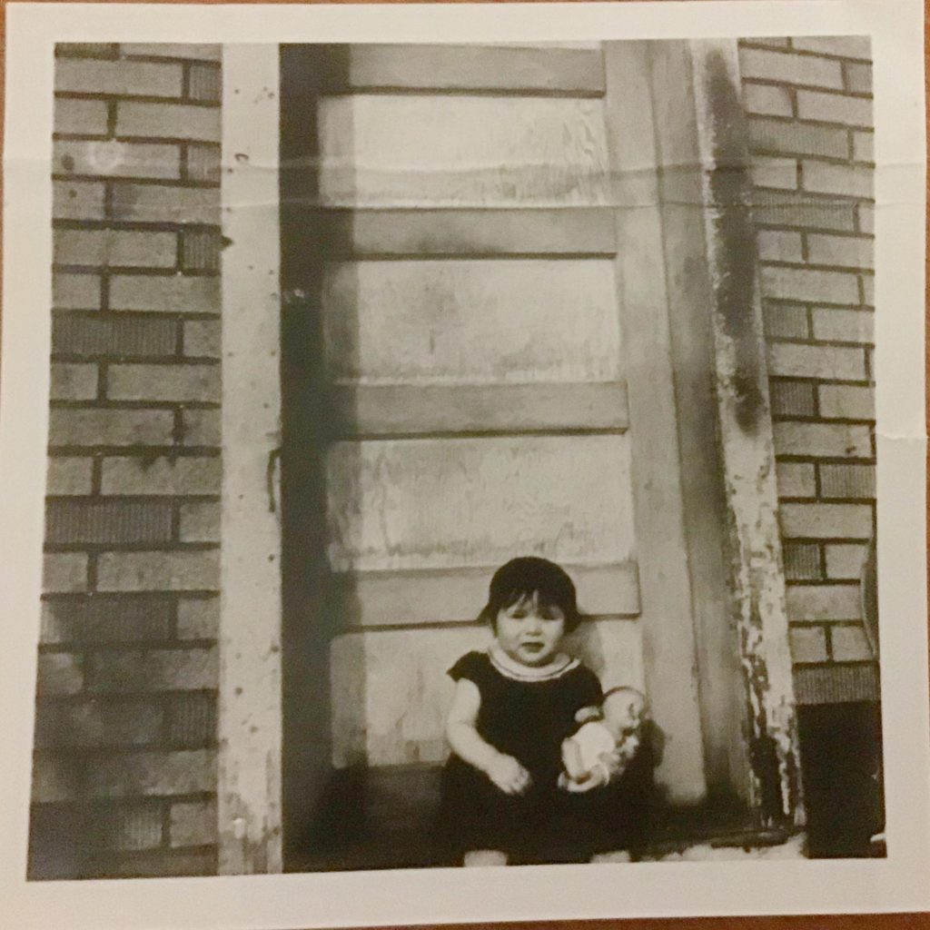 A black and white image of Mary Ann Assinabe as a baby, sitting in the doorframe of a building and holding a baby doll in her arms.