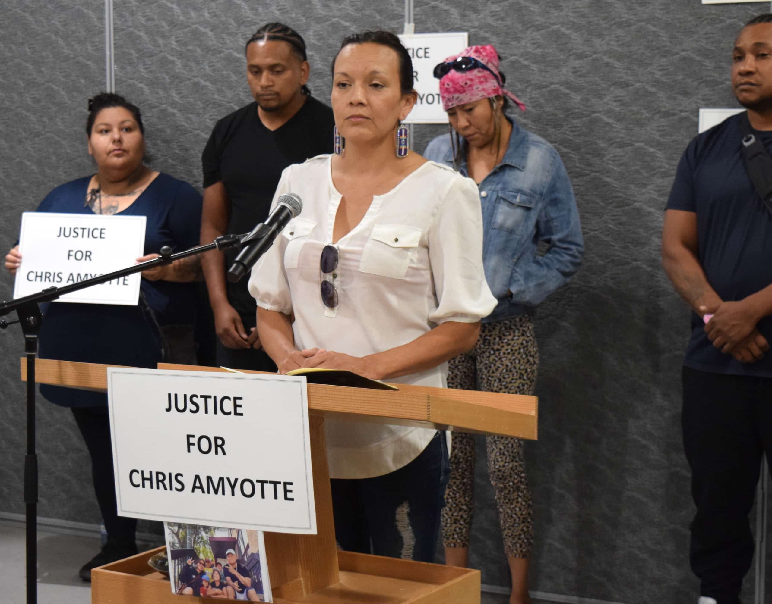 A woman stands at a podium with a sign in front of her that reads "Justice for Chris Amyotte"