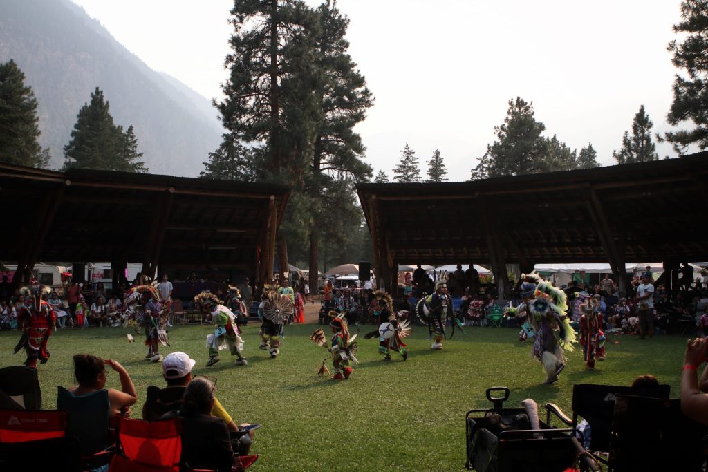 Around nine dancers in their regalia are dancing on a clearing of grass within a wooden arbor. There are crowds of spectators in a circle around the clearing.