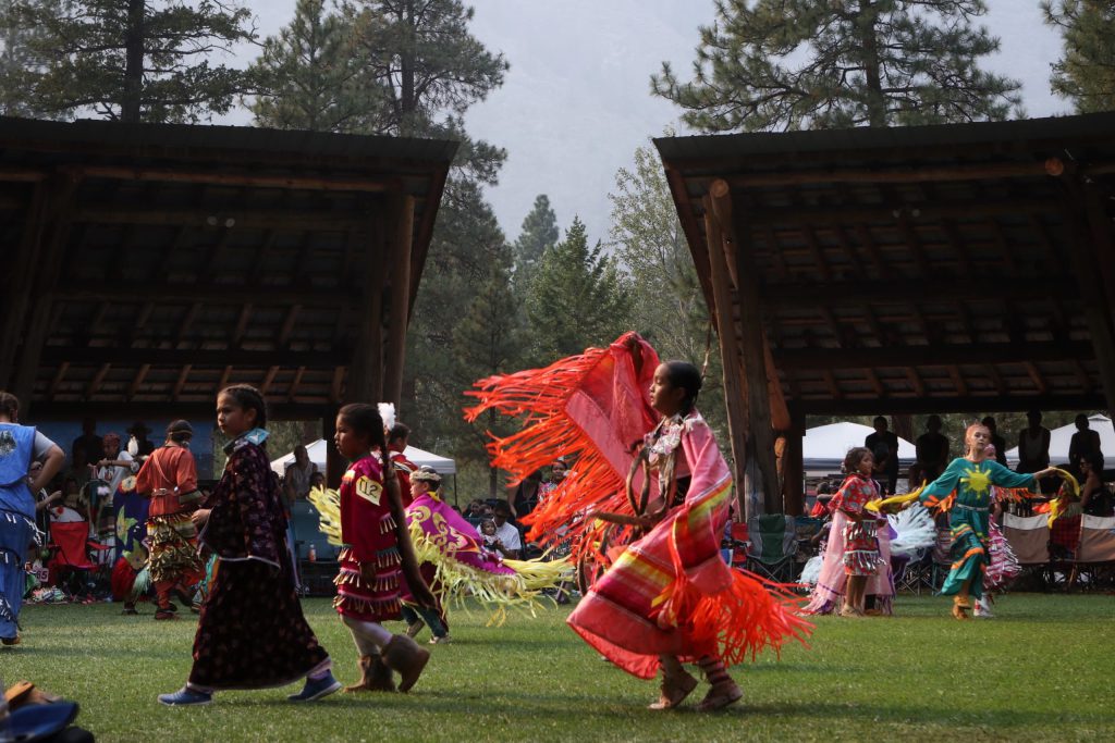 Five kids are pictured in their regalia, two are wearing patterned dresses, two are in a jingle dress, and another a pink dress with matching shawl. They are dancing in the grassy clearing in the centre of the arbor.