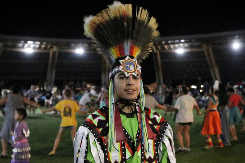 A young man stands in his regalia including a hair roach headdress as people dance in the powwow grounds behind him