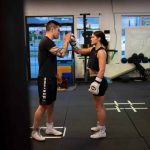 An Indigenous man stands face to face with a younger woman, both dressed in black shirts and shorts with white sneakers. The young woman is wearing white boxing gloves, and is punching her right glove into the man's right palm during a training session in a boxing gym in downtown Snuneymnuxw.