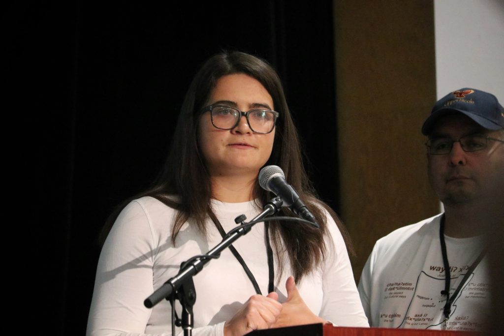 An Indigenous woman wearing white shirt and glasses stands speaking to forum about sylix conservation knowledge.