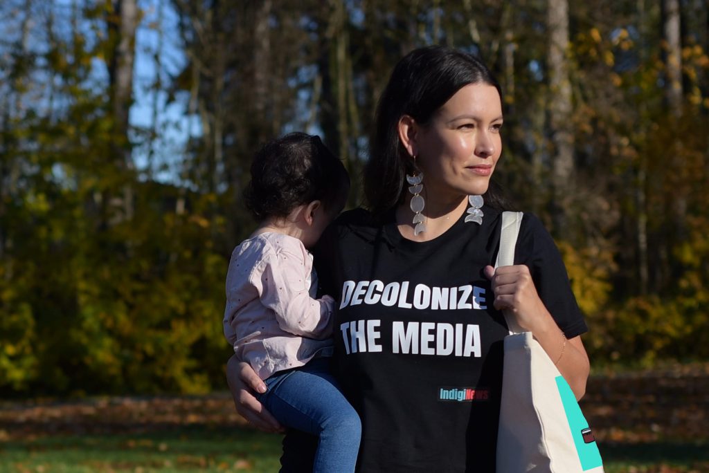Anna McKenzie poses in an IndigiNews t-shirt that has the words, "Decolonize the media" on it while holding her child, whose face is turned away.
