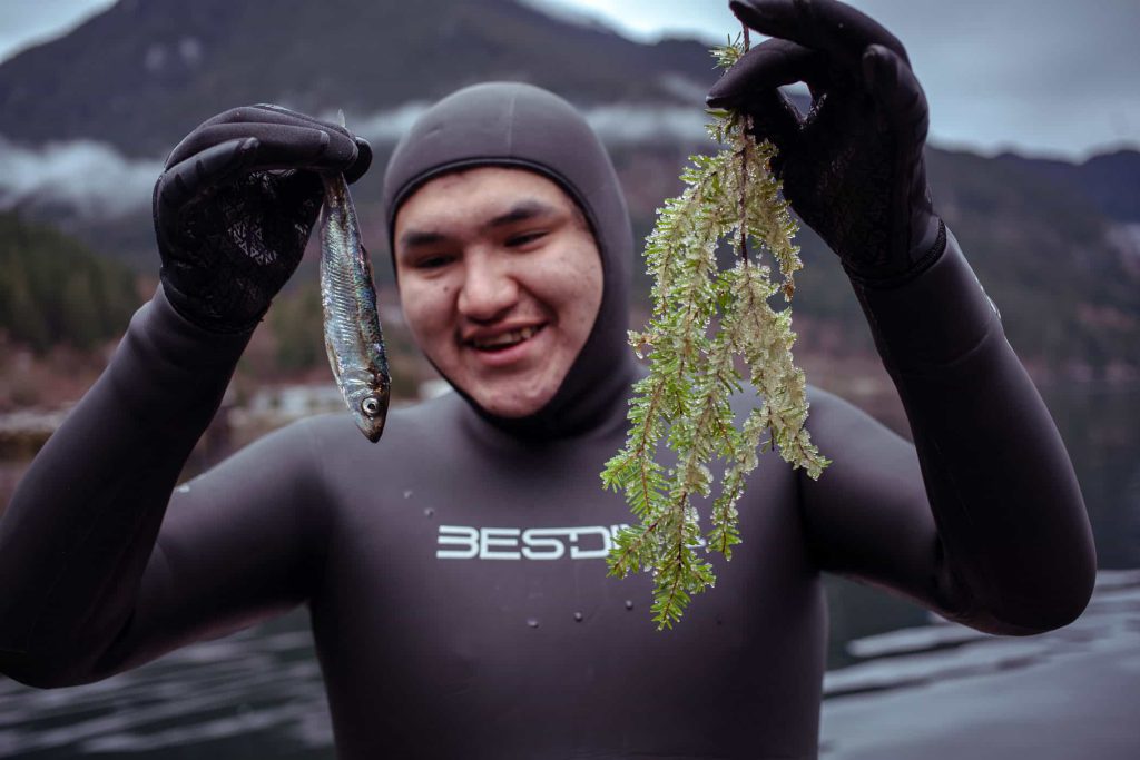 A young man in a wetsuit holds up a herring he found floating along the shore and a hemlock sprig laden with herring eggs