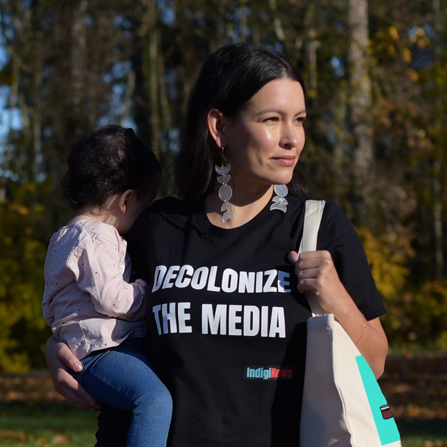 Anna McKenzie poses in an IndigiNews t-shirt that reads, "Decolonize the media," while holding a child whose face is turned away.