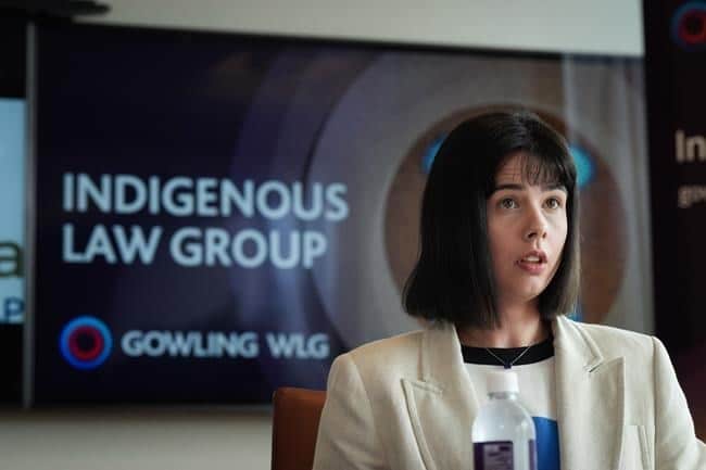 A dark-haired woman in a white blazer stands in front of a screen that reads, "Indigenous Law Group."