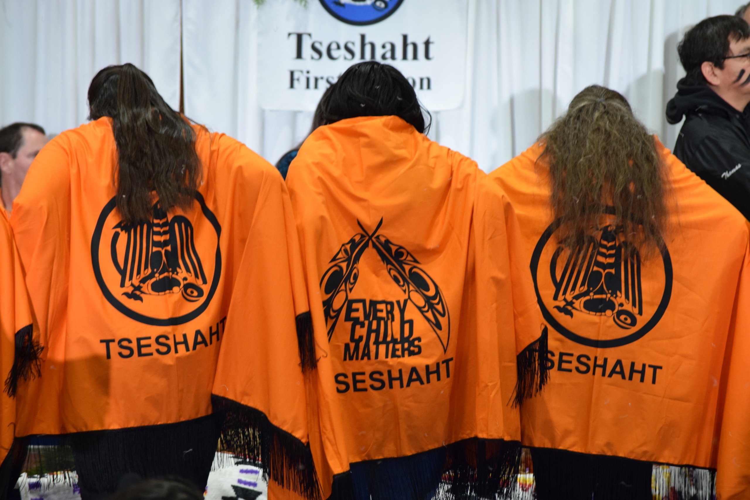 Three women stand with their backs to the camera, and they each wear orange capes with the Tseshaht and Every Child Matters logos