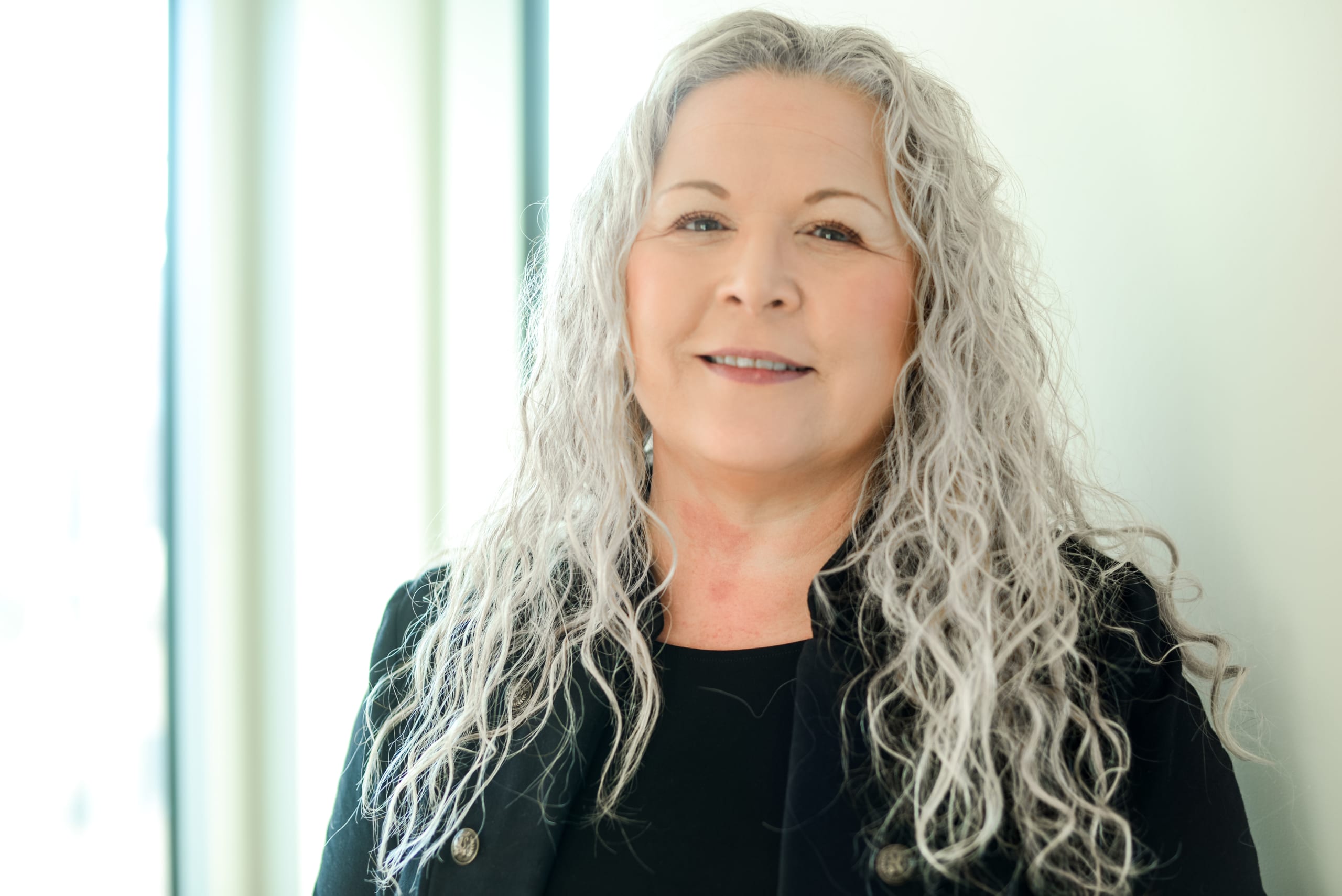 An Indigenous woman with long, curly silver hair smiles at the camera in a black top.