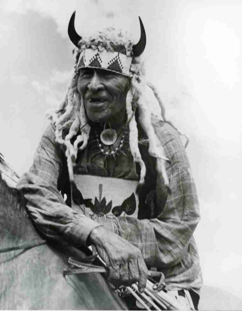 A black and white photo of an old Cree man named Fine Day, sitting on a horse wearing a beaded headdress with horns on it.
