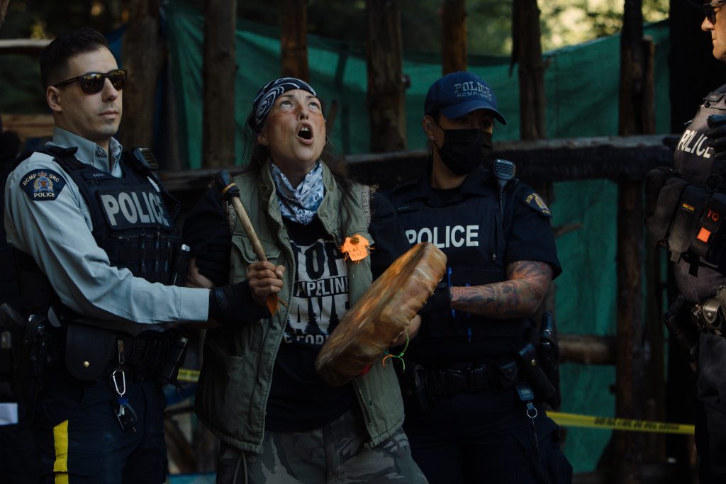 Uncle Ricco continues to sing and drum as RCMP CIRG officers arrest them at Savage Patch, a camp blockading old growth logging, in Pacheedaht territory near “Port Renfrew” on “Vancouver Island” on Aug. 15, 2023. Three people were arrested and released without charges. Photo by Amber Bracken 