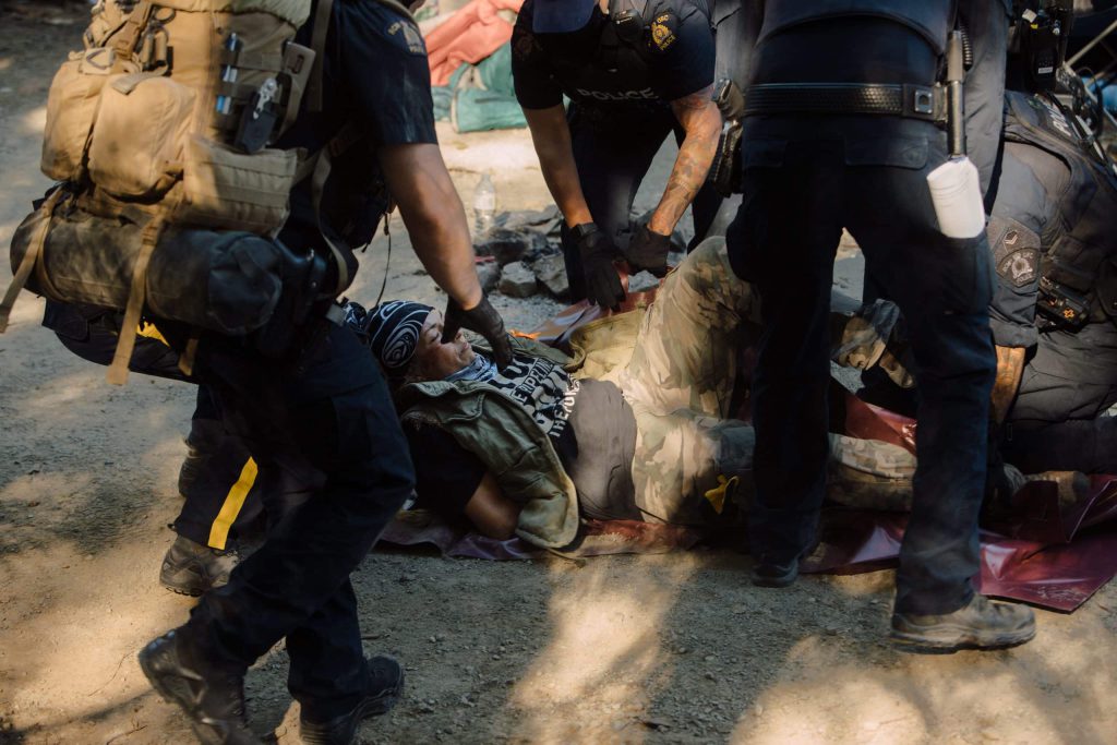 Uncle Rico is laid on her back during her arrest by RCMP CIRG officers at Savage Patch, a camp blockading old growth logging, in Pacheedaht territory near “Port Renfrew” on “Vancouver Island” on Aug. 15, 2023. At least three people were arrested. Photo by Amber Bracken 