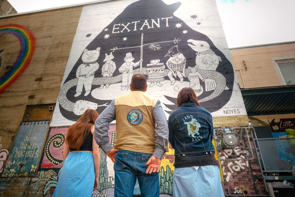 Aaron Fitzpatrick and his daughters look up at a mural painted by Sinixt artist Emma Noyes in an alleyway in ki’yamələp place of dead trees (Nelson), June 2023. Photo by Mike Graeme