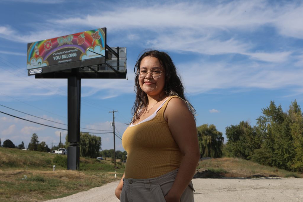 Sarah Jones, a Two-Spirit Anishinaabe freelance artist from Treaty 3 Shoal Lake 40, stands near the “You Belong, kʷu yʕayʕát kʷuʔ čn̓k̓ʷɬn̓xiʔm̓ (We all join in together / We are all a part of it together)” billboard that she designed in syilx homelands on Sept. 7. Photo by Aaron Hemens