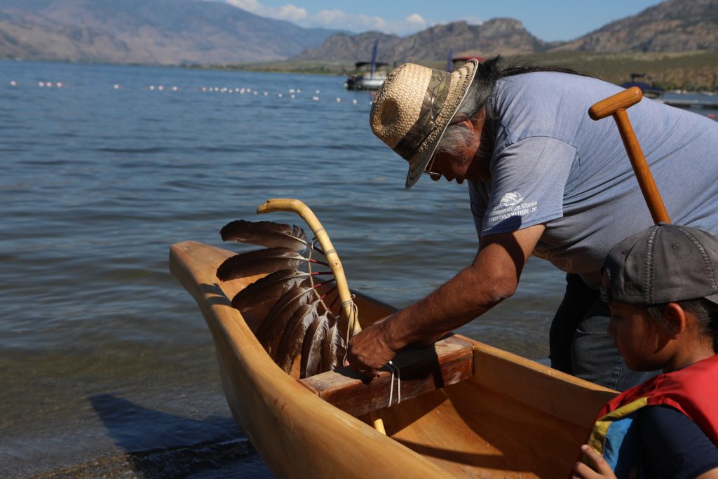 Herman Edward ties an eagle staff to a canoe, prior to the launch of the 2024 Sukʷnaqin canoe journey. Photo by Aaron Hemens
