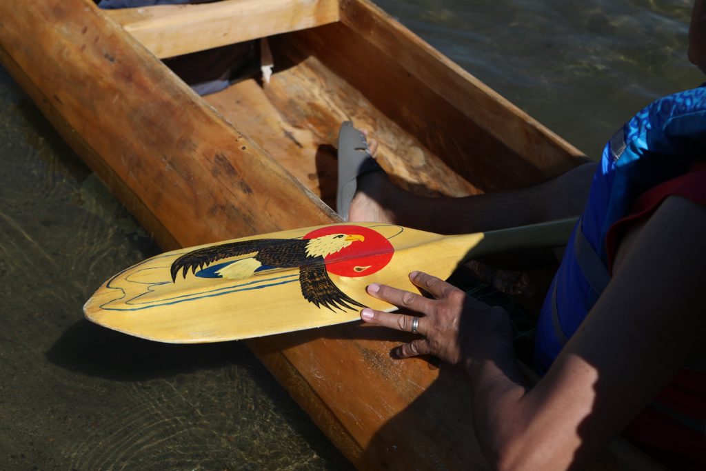 A community member readies their paddle during the launch of Herman Edward ties an eagle staff to a canoe, prior to the launch of the 2024 Sukʷnaqin canoe journey. Photo by Aaron Hemens
