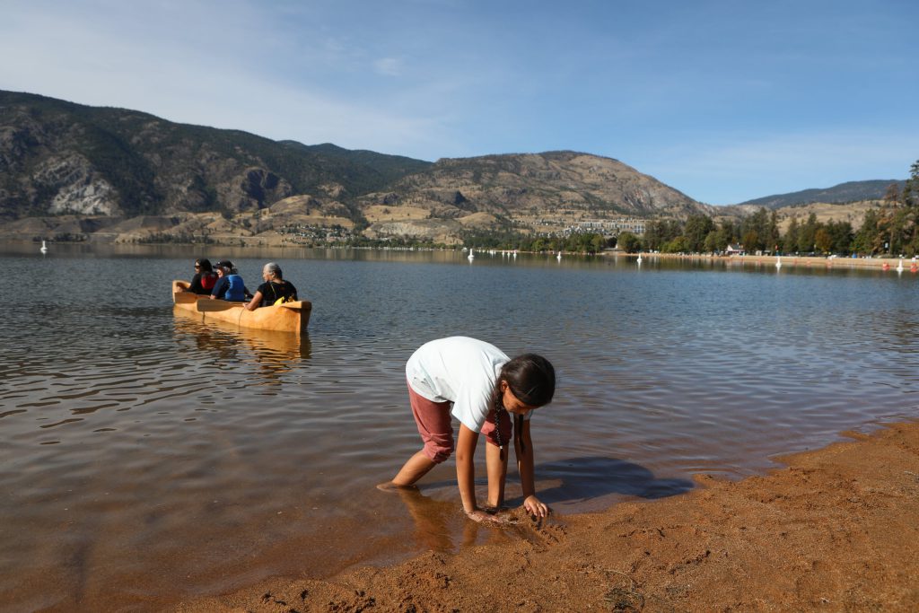 A child plays in the sand on the shores of Skaha Lake, as paddlers in a canoe make their way to sx̌ʷəx̌ʷnitkʷ (Okanagan Falls) on Sept. 21. Photo by Aaron Hemens