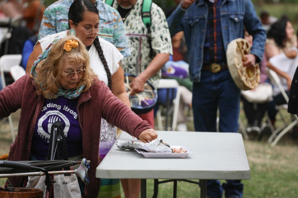 Community members line up and take pieces of salmon during the syilx Nation’s 2024 salmon feast and celebration at sx̌ʷəx̌ʷnitkʷ (Okanagan Falls) on Sept. 22. Photo by Aaron Hemens