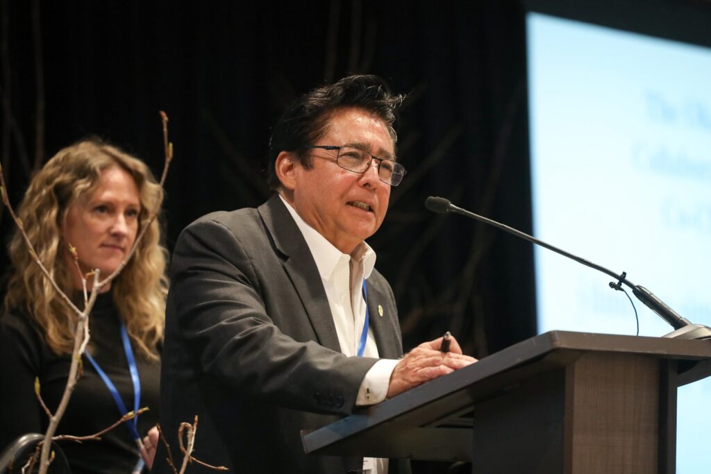 Chief simo Robert Louie of Westbank First Nation, co-chair of the Okanagan-Similkameen Collaborative Leadership Table, speaks at the Okanagan Nation Alliance's 2026 siwɬkʷ (Water) and Climate Forum on March 23 in Kelowna, B.C. Behind him is fellow co-chair Tricia Brett, councillor for the District of Lake Country. Photo by Aaron Hemens