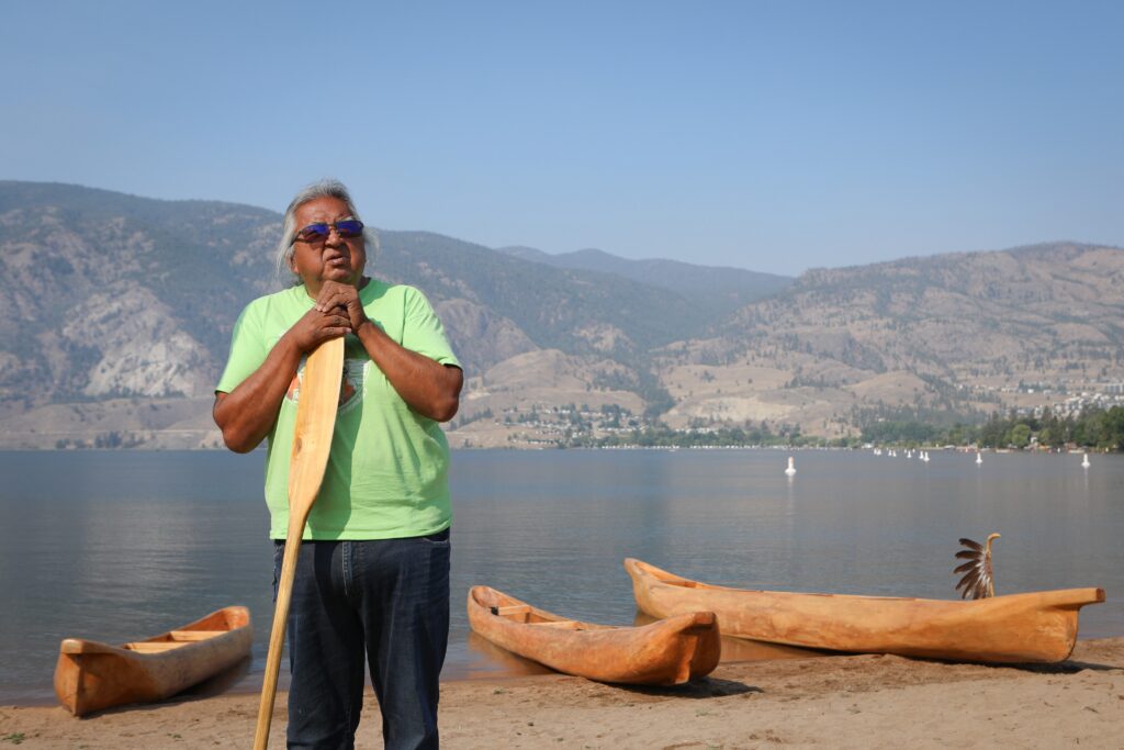 Herman Edward stands at Skaha Lake on July 31, near three dugout canoes that he carved. The canoes were used in the syilx Nation’s 2026 Unity Trek Commemorative Paddle. Photo by Aaron Hemens